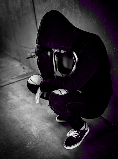 A person wearing a hooded jacket and ripped jeans squats in a dimly lit, shadowy environment, resting their head on their hand, reflecting the weight of depression.