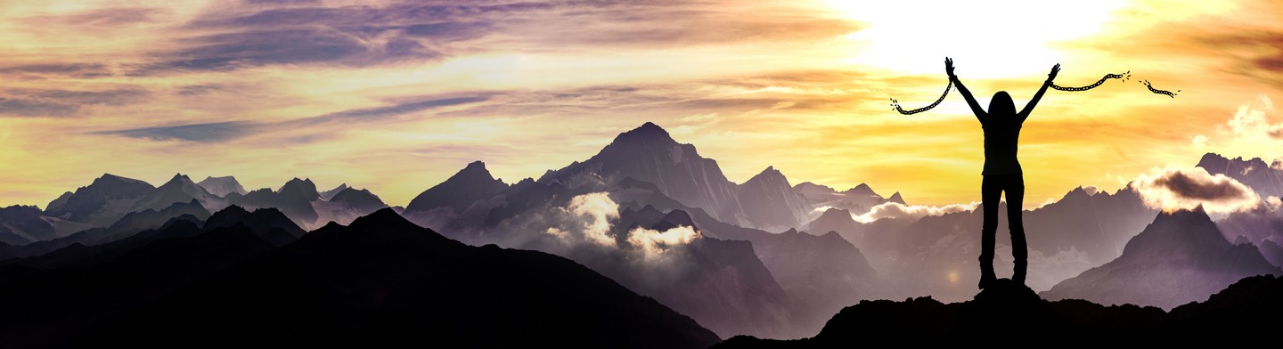 Silhouette of a person with broken chains in their hands standing atop a mountain, facing a sunrise or sunset with a panoramic view of mountain peaks and clouds—a powerful symbol for narcissistic abuse recovery.