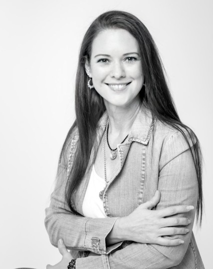 Black and white photo for the 'About Me' section: A person with long hair, wearing a light-colored jacket and a white shirt, smiling and sitting with their arms crossed.